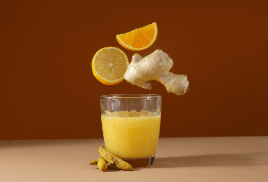 A glass of yellow liquid with a lemon slice, an orange slice, and a piece of ginger floating above it