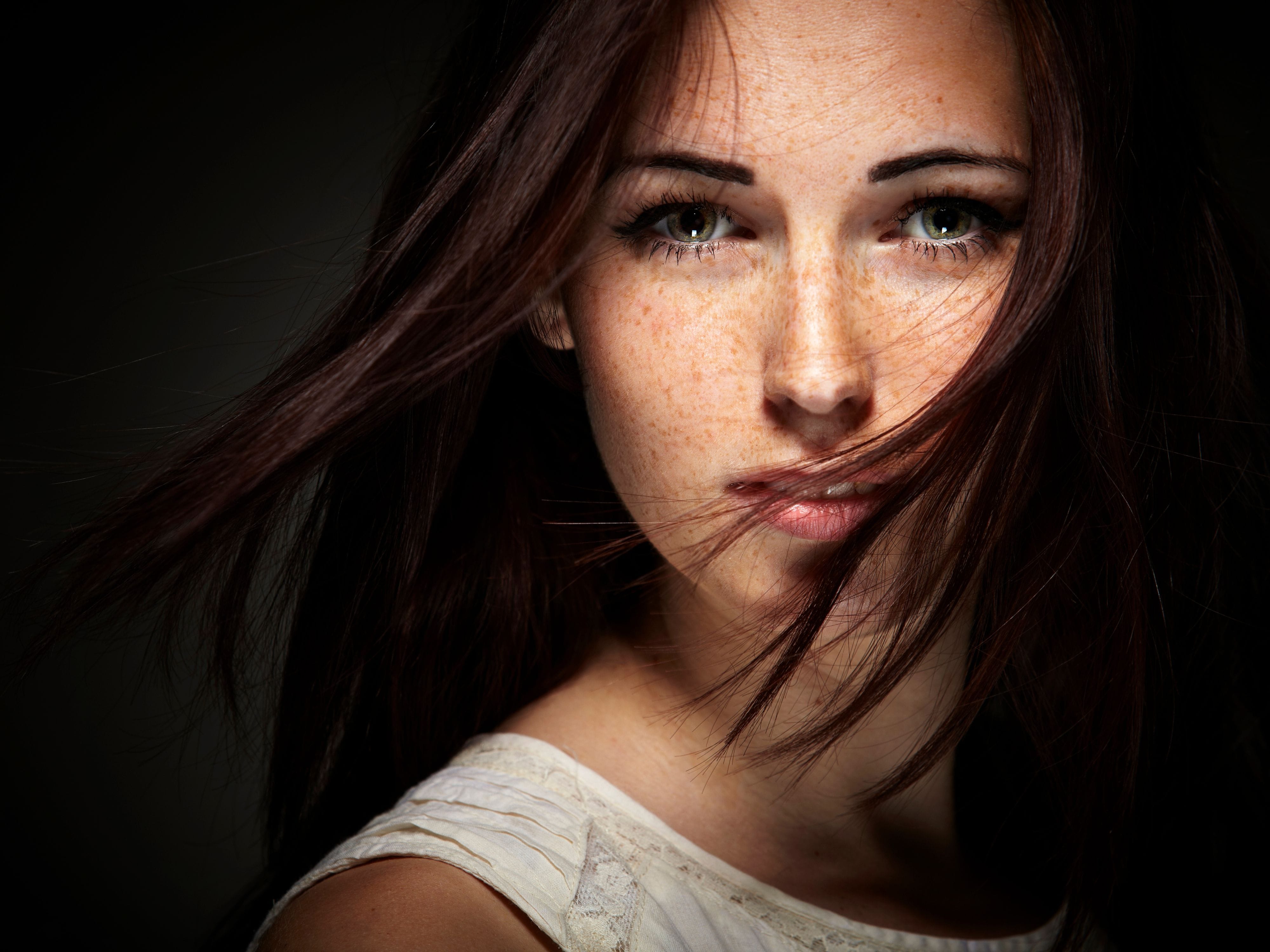 A portrait of a woman with green eyes and freckles, her dark hair blowing across her face