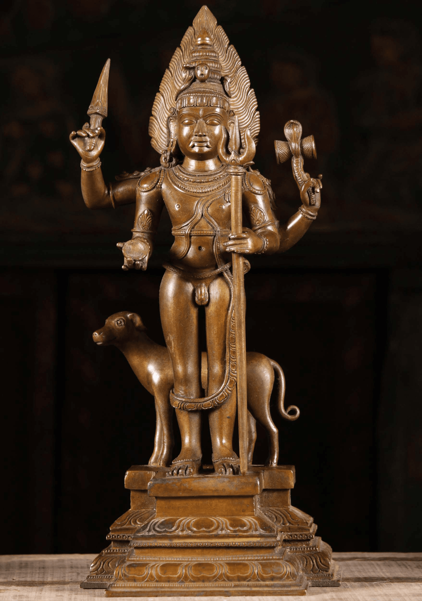 This photo of a bronze idol of Lord Kala Bhairava shows His detailed iconography, including a dog standing sideways, right behind Him. In His 4 hands, holds a Trishul, Damaru wrapped with a snake, Kapala, and Pasha.